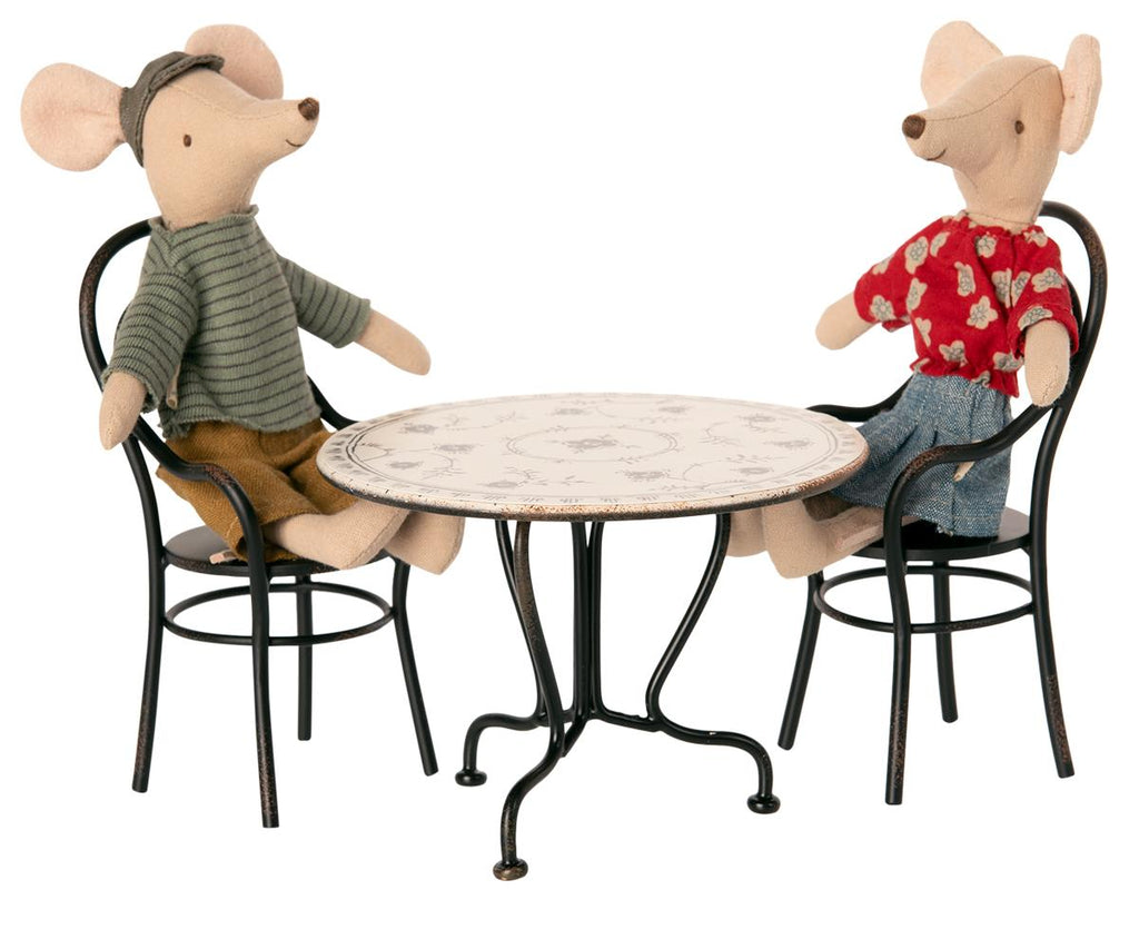 Two fabric mice dolls sit across from each other at a small round Maileg Bistro Table With Chairs with a decorative top. One mouse wears a green striped shirt and cap, while the other sports a red flower-patterned shirt and jeans. Both mice are seated on black metal chairs, ready for their Sunday supper.