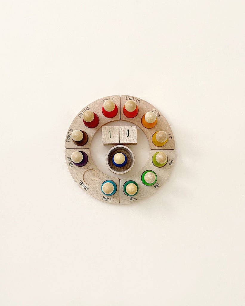 An antique rotary telephone dial, repurposed as a decorative piece, showcasing colorful buttons from a Grapat Perpetual Calendar with Peg Figures labeled with days of the week within each number slot, against a plain background.