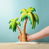 A hand arranging the Set of Handmade Wooden Palm Trees & Waves on a tabletop, against a blue background. The trees have detailed trunks and vibrant green leaves.