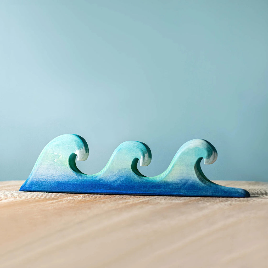 Three Handmade Wooden Palm Trees & Waves sculptures, painted with a gradient from navy blue to teal using non-toxic paint, are aligned on a wooden surface against a light blue background.