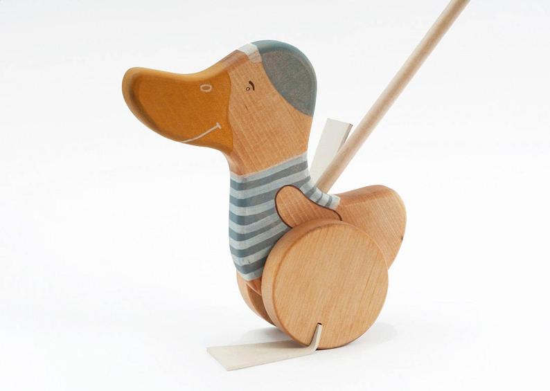 A Handmade Wooden Duck Push Toy crafted from sustainably harvested birch wood in the shape of a cartoon-style dog on wheels, painted in stripes of grey and beige, with a stick attached to its back for pushing.