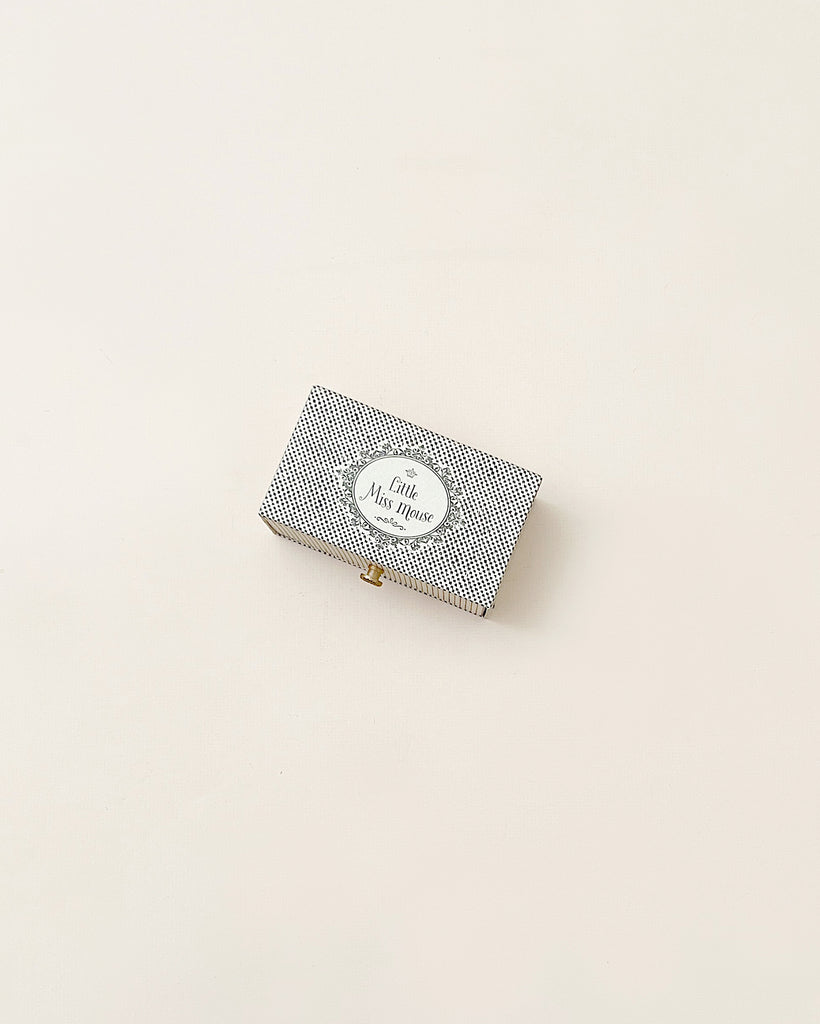 A small, elegant Maileg Dancing Mouse in Daybed with a houndstooth pattern and a golden clasp. It includes a label saying "la vie est belle Paris" on a beige background.
