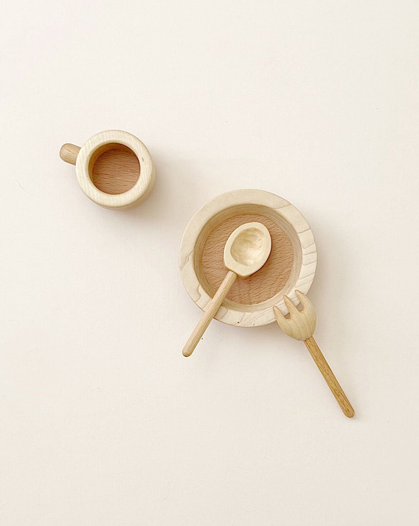 A minimalist Handmade Pretend Dinnerware Set consisting of a small cup with a drink, a plate with a bowl and handmade wooden cutlery, all arranged neatly on a light beige surface.