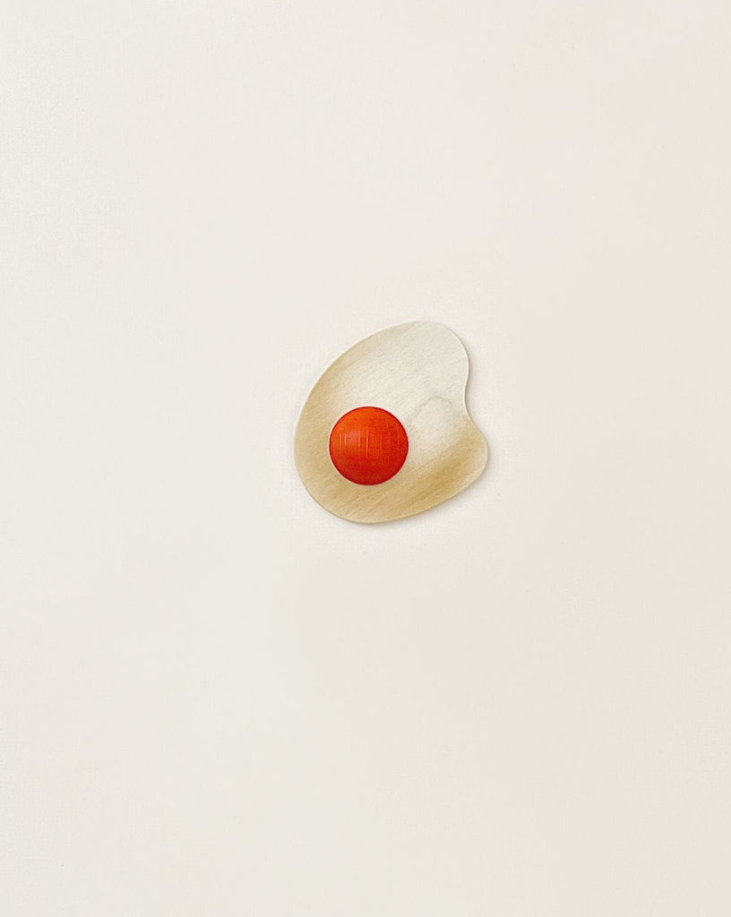 A small, irregularly shaped white object with a smooth texture is centered on a plain, light background. It has a bright red circular indentation in its middle, resembling a minimalist artistic interpretation of an Erzi Wooden Sunny-Side Up Egg, and looks like it could be part of an exquisitely handcrafted toy made from solid beech wood.
