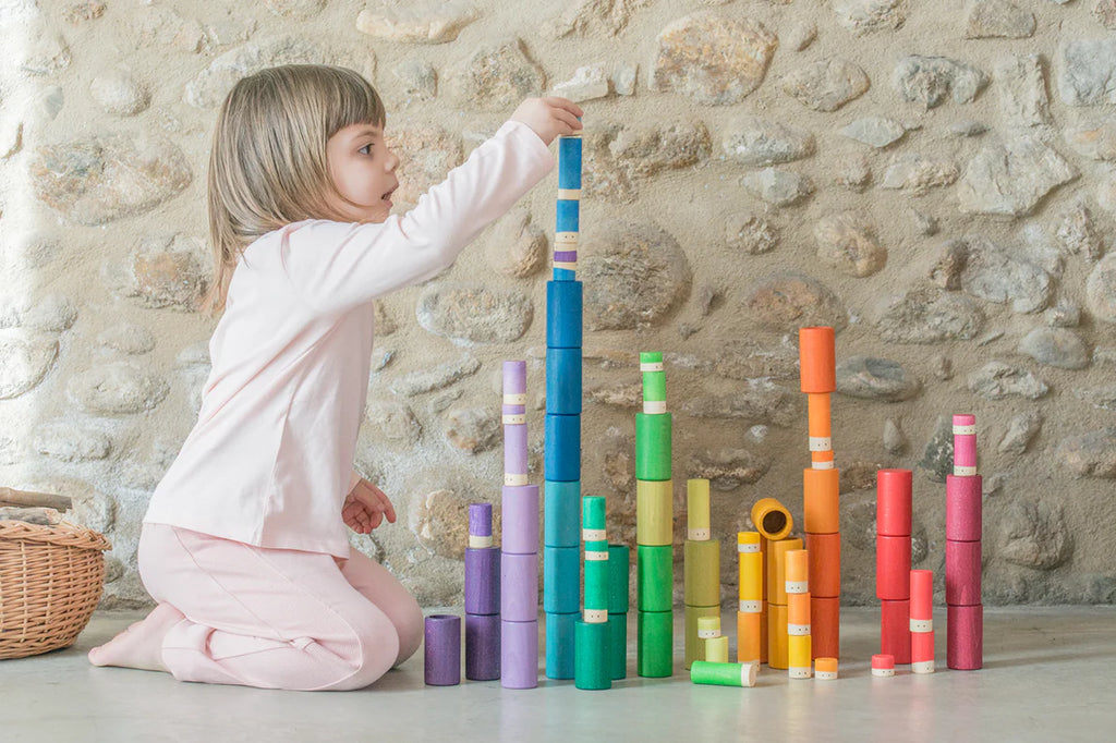 A young child carefully places a Grapat Lola Stack and Sort Play Set 72 pcs atop a colorful stack of similar tubes, engaging in imaginative play while creating a tall tower. They kneel on the floor in a room with a stone wall and a.
