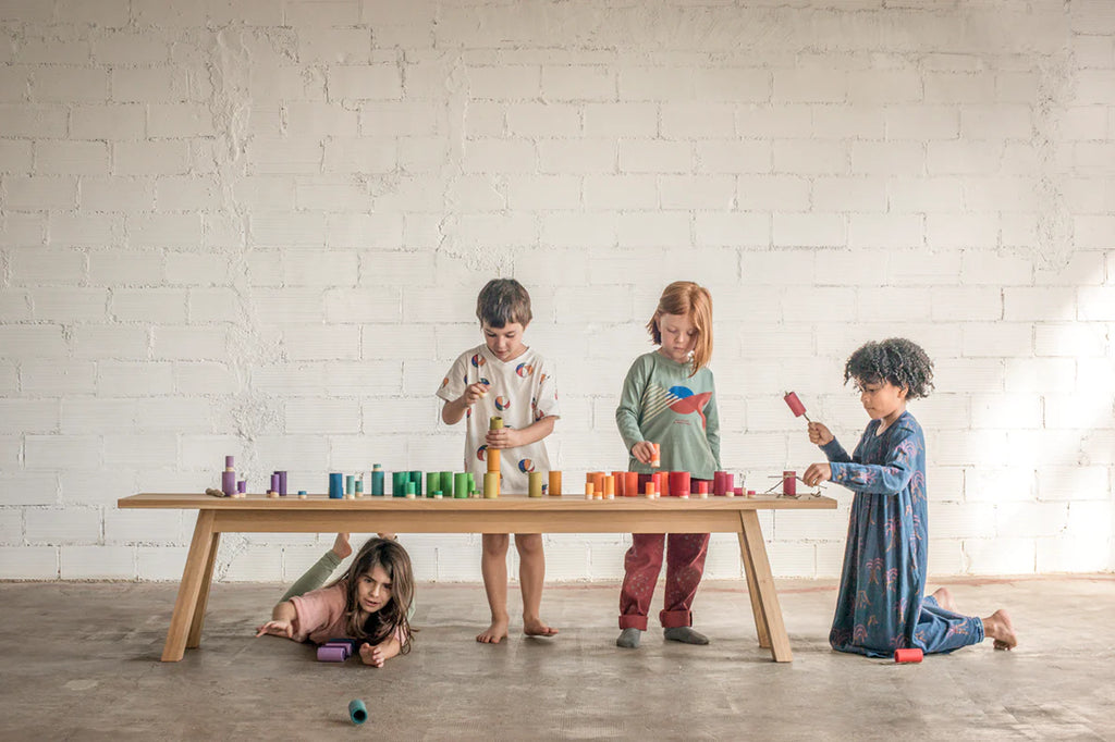 Four children engage in imaginative play with colorful Grapat Lola Stack and Sort Play Set 72 pcs and musical instruments on and around a wooden table in a room with white brick walls.