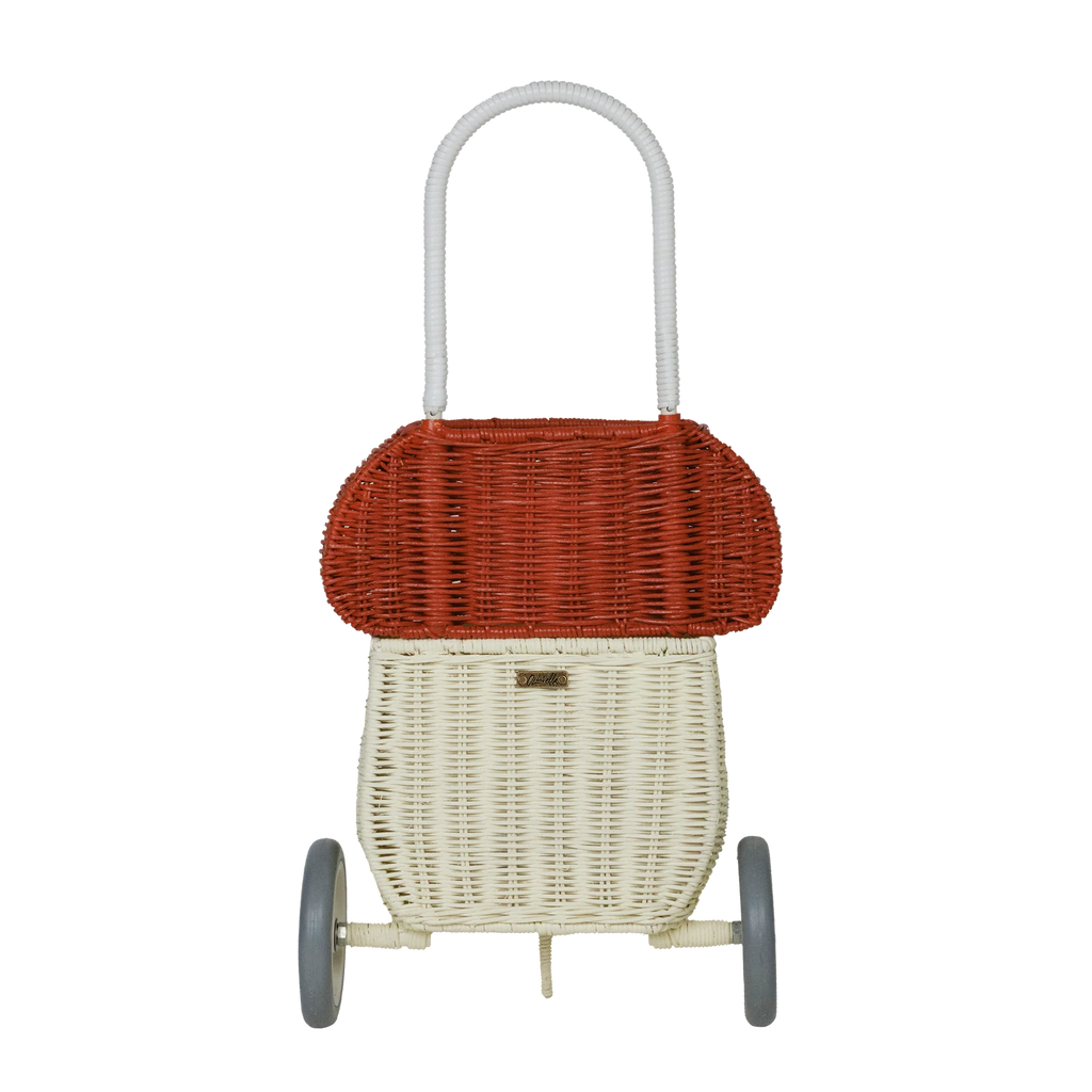 A vintage two-wheeled wicker shopping trolley, whimsically dubbed the Olli Ella Rattan Mushroom Luggy - Red, features a red top and cream-colored body, standing in front of a black background with white horizontal lines.