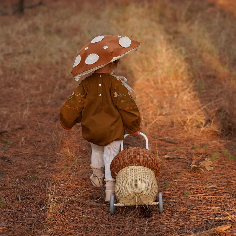 A child dressed as a mushroom, with a polka-dotted cap, pulls an Olli Ella Rattan Mushroom Luggy - Natural through a forest covered with brown pine needles.