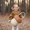 A joyful toddler with blonde hair, wearing a brown blouse and white pants, laughs while holding an Olli Ella Rattan Mushroom Luggy - Natural basket in a forest during autumn.