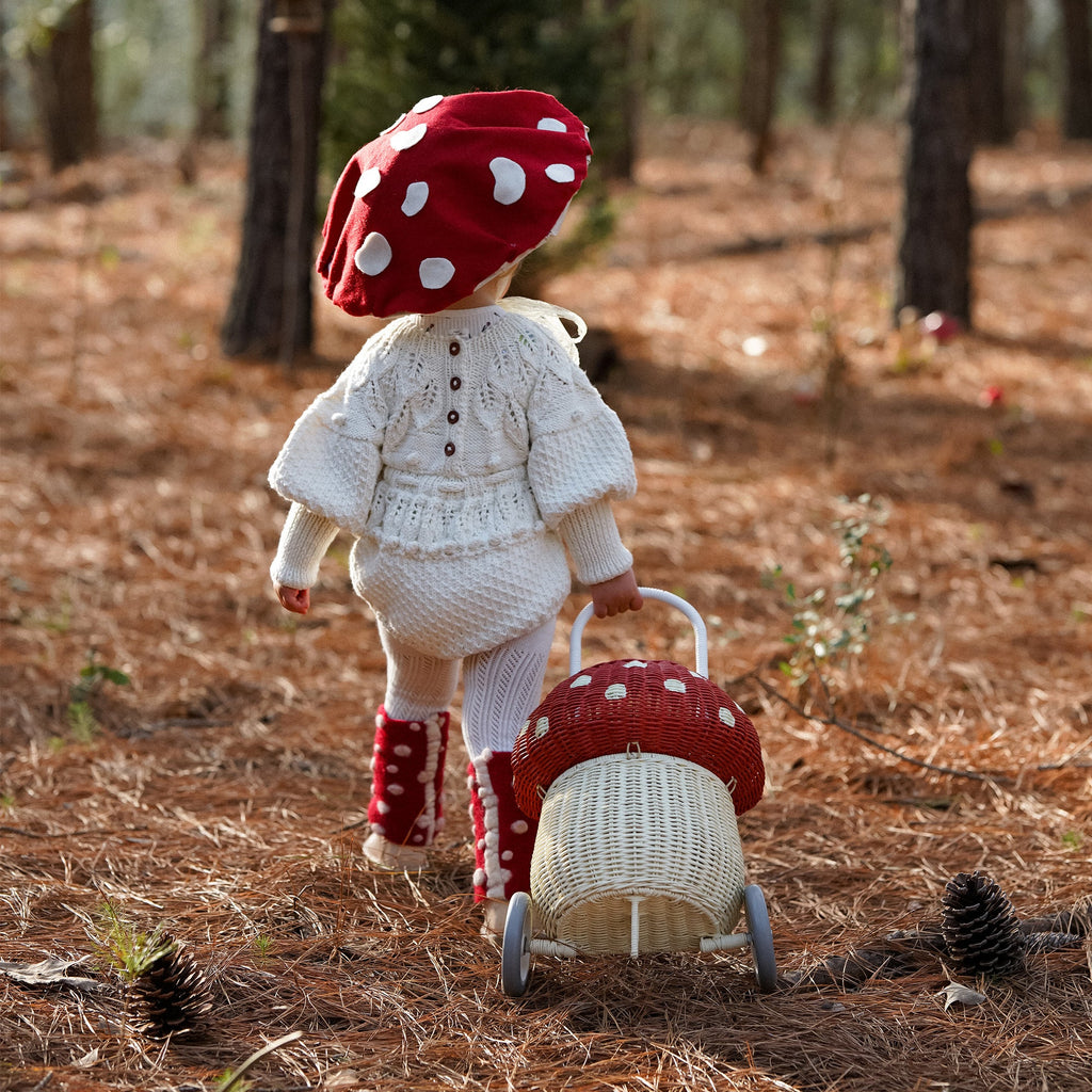 A doll with a red polka-dot mushroom cap head and a knitted white outfit pushes a small hand-woven Olli Ella Rattan Mushroom Luggy - Red stroller through a pine forest, surrounded by pine cones on the ground.