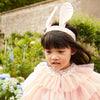 A young girl wearing a Meri Meri Peach Tulle Bunny Costume, including ears headband and a ruffled peach dress adorned with daisies, playing in a garden with lush greenery and flowers in the background.