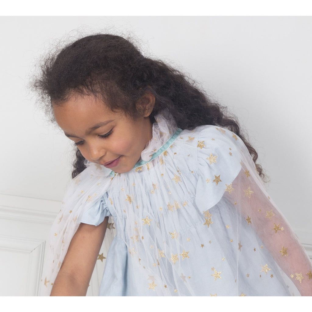 A young girl with curly hair, smiling and looking down, wearing a Meri Meri White Tulle Star Cape Costume adorned with gold glitter stars.