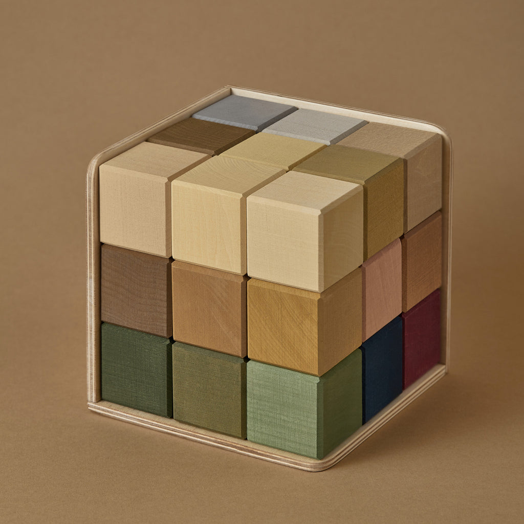 A neatly assembled Raduga Grez | Small Cube Blocks, consisting of smaller blocks in muted shades of brown, green, and gray—all painted with non-toxic paint—contained within a transparent acrylic case against a tan background.