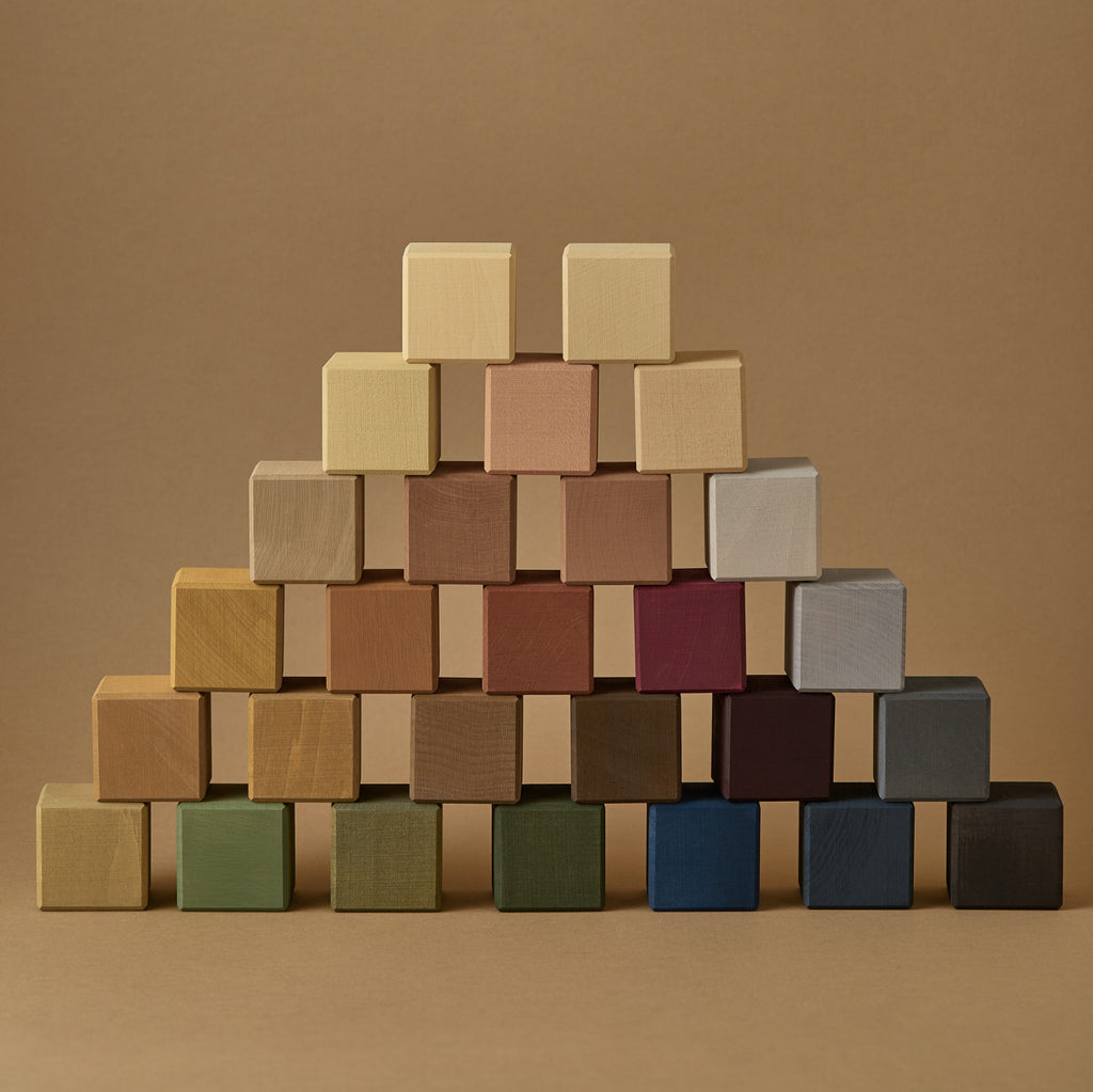 A neatly arranged pyramid of Raduga Grez Small Cube Blocks, coated in non-toxic paint, in various shades, set against a soft brown background. The arrangement showcases a gradient of colors from natural wood tones to deeper.