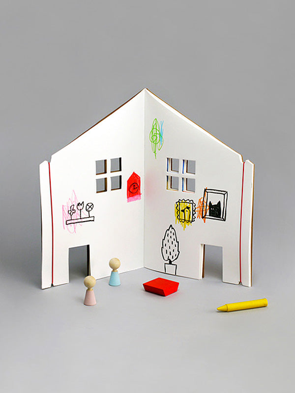 A simple white cardboard Dollhouse Drawing Book with child-like drawings on its walls, featuring small windows. In front, there are two teddy bears and crayons.