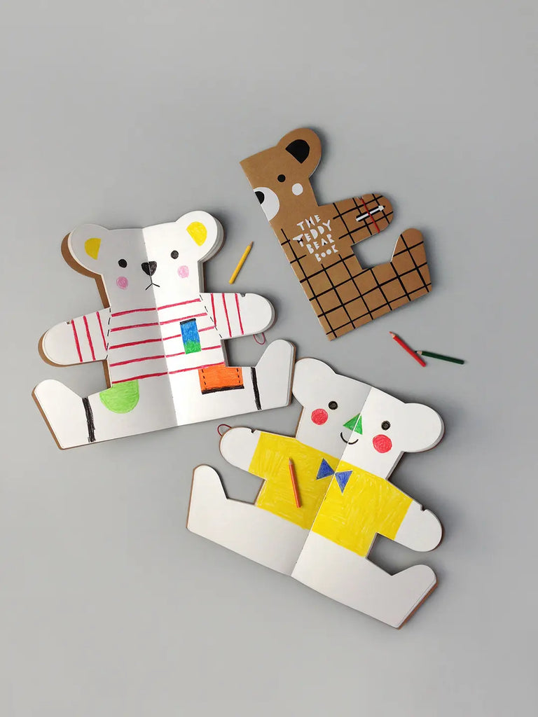 Three cardboard Teddy Bear Drawing Books lying on a gray surface, each book uniquely decorated with colorful geometric designs and placed next to crayons.