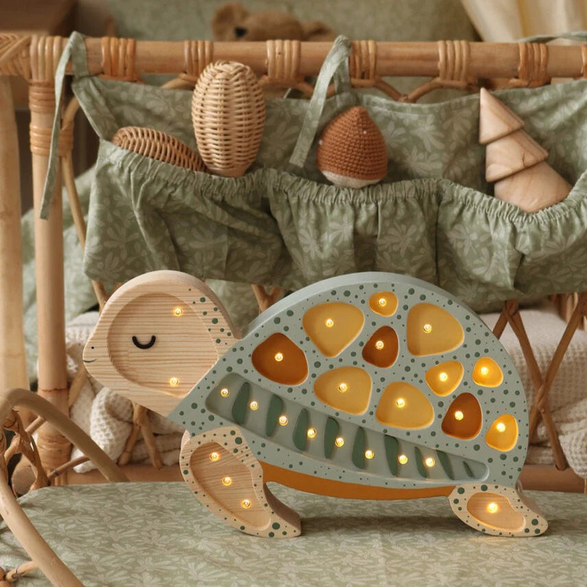 A cozy wicker crib is adorned with a hanging, double-decked holder filled with wicker and wooden toys. In the foreground, a children's night light shaped like a Little Lights Turtle Lamp emits a gentle glow from its cut-out patterns, resting on a decorative cushion with a leafy design.