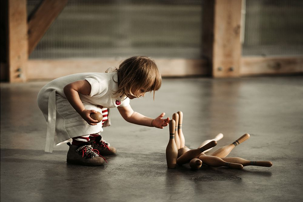 Child wearing white dress and brown boots holding a wooden bowling ball. 3 standing wooden bowling pins standing and 7 wooden bowling pins laying.