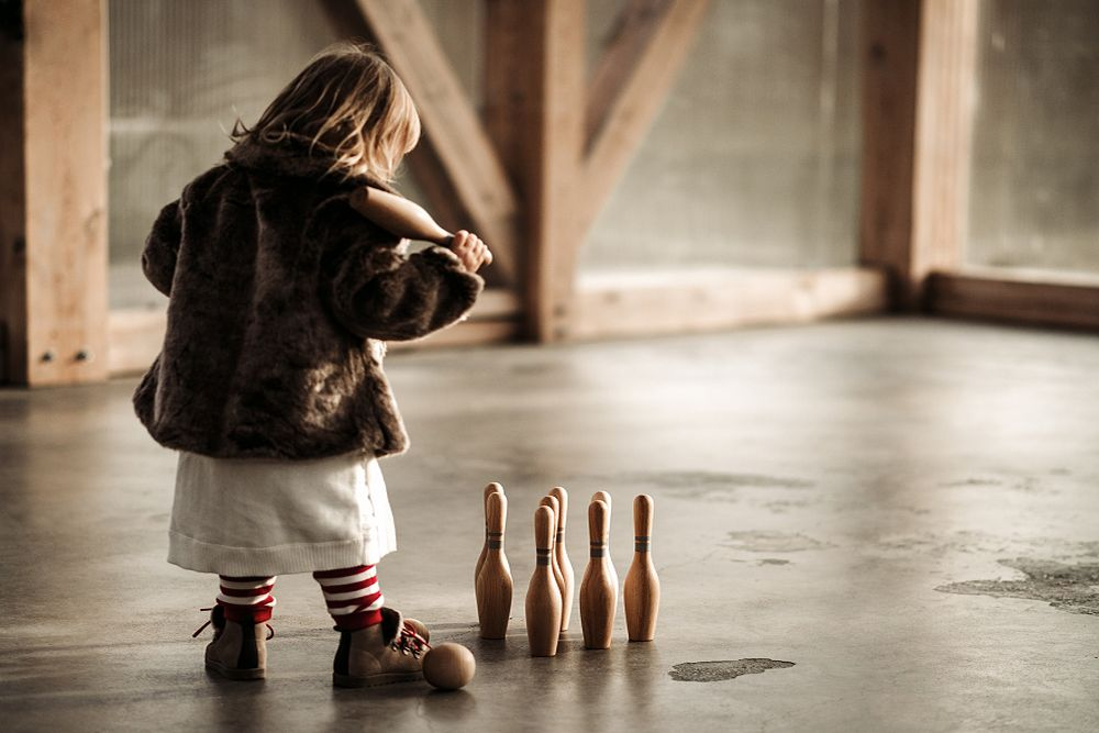 Child wearing white dress, brown jacket and brown boots. Child holding one wooden bowling pin. 8 wooden bowling pins and one wooden bowling ball on the floor next to child.