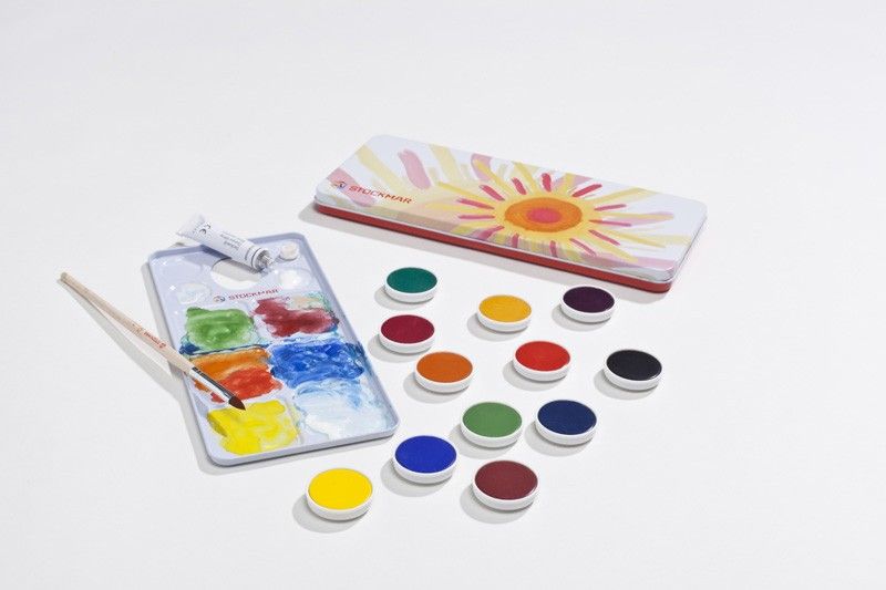 A set of Stockmar Opaque Colors - 12 Colors watercolor paints with various colors open next to a palette and paintbrush, all against a plain, light background. A tin with a sunflower design is partially visible.