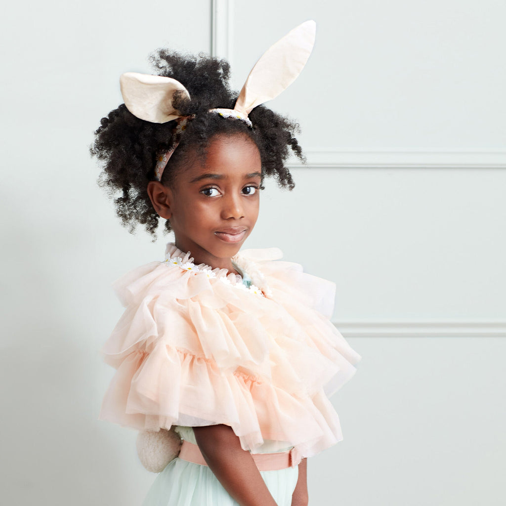 A young girl with curly hair wearing a Meri Meri Peach Tulle Bunny Costume, looking towards the camera with a gentle smile, standing against a light grey background.