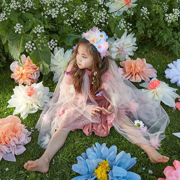 A young girl in a pink floral dress and Meri Meri Flower Cape sits among oversized colorful tulle flowers on a lush green lawn.