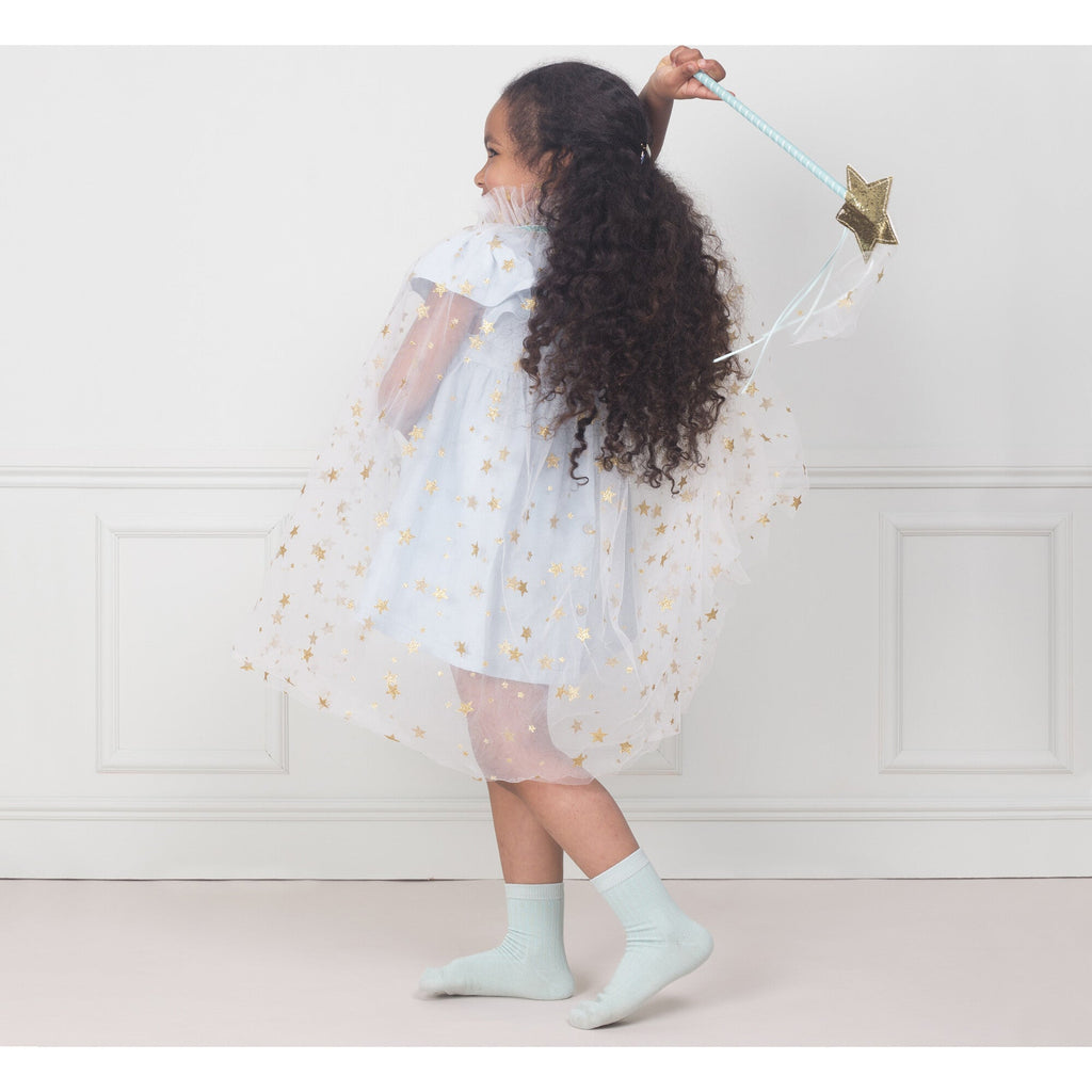 A young girl with long curly hair wearing a Meri Meri White Tulle Star Cape Costume and holding a sparkly star wand, playfully twirls in a well-lit room with a subtle wall.
