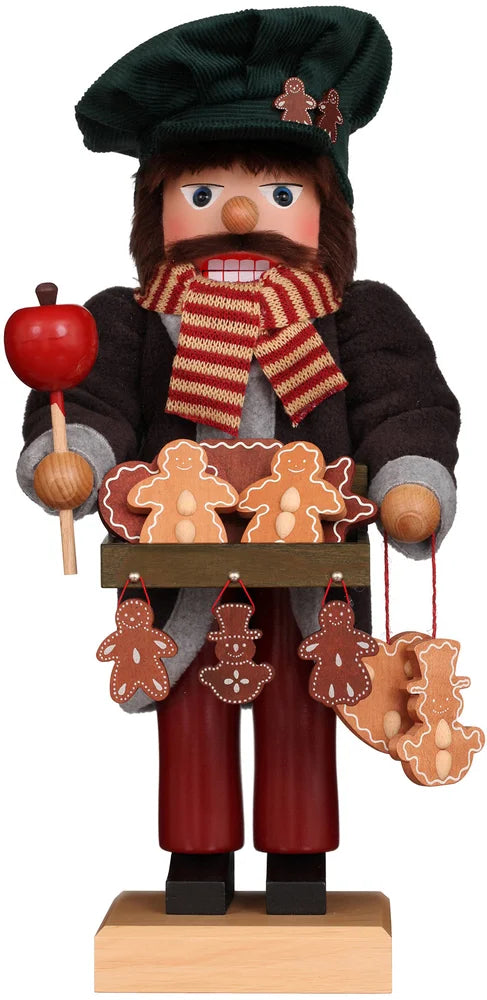 The Christian Ulbricht Collectible Wooden Nutcracker - Gingerbread Vendor Limited Edition 1000 wears a green hat, red-striped scarf, dark coat, and holds a candied apple plus a tray of gingerbread people cookies with more hanging below.