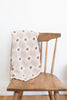 A wooden chair displays the Organic Cotton Baby Blanket - Teddy, in light beige with a diamond pattern and small brown floral designs, draped over the backrest. Made in Portugal, this soft baby blanket is set against a plain white wall.