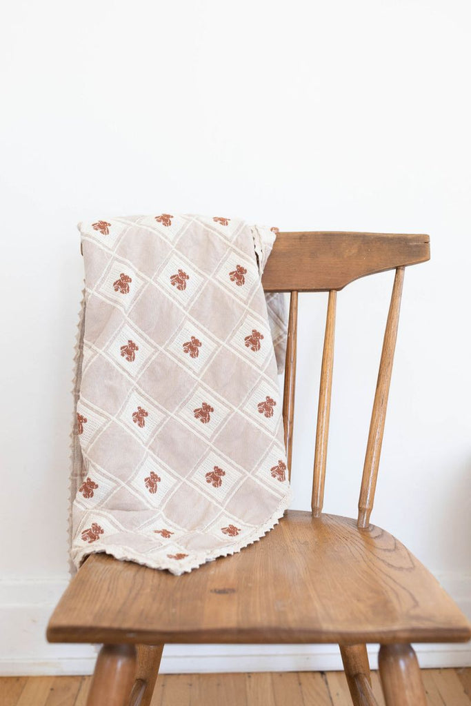 A wooden chair displays the Organic Cotton Baby Blanket - Teddy, in light beige with a diamond pattern and small brown floral designs, draped over the backrest. Made in Portugal, this soft baby blanket is set against a plain white wall.