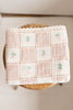 The Organic Cotton Baby Blanket - Fleur, a pastel pink blanket with a grid pattern and small embroidered cherries made in Portugal, is displayed on a round wooden stool.