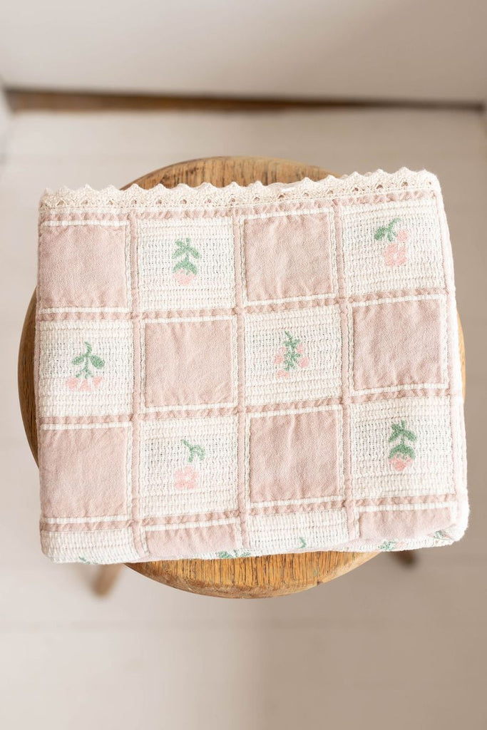 The Organic Cotton Baby Blanket - Fleur, a pastel pink blanket with a grid pattern and small embroidered cherries made in Portugal, is displayed on a round wooden stool.