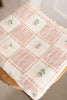 The Organic Cotton Baby Blanket - Fleur features a pink and cream patchwork grid with small floral details, draped over a wooden surface. Made in Portugal.