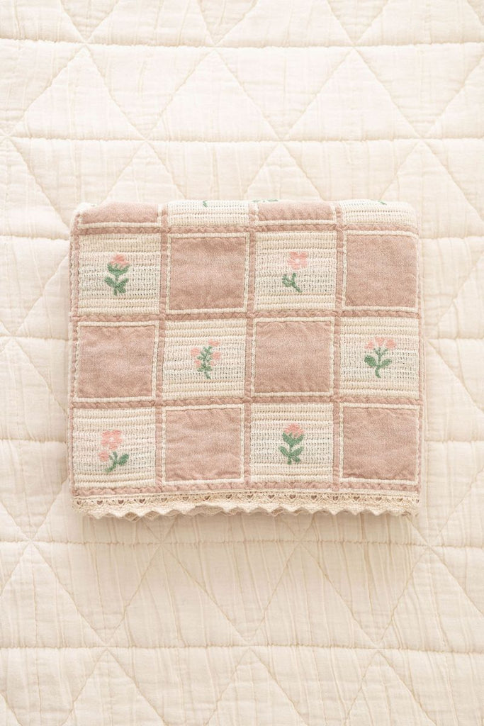 The Organic Cotton Baby Blanket - Fleur features a patchwork design with pink and green embroidered flowers, made in Portugal from organic cotton, shown on a cream quilted surface.