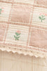 A close-up of the Organic Cotton Baby Blanket - Fleur shows its checkered pattern, pink embroidered flowers, and lace-trimmed edge. Made in Portugal from soft, vintage-style fabric.