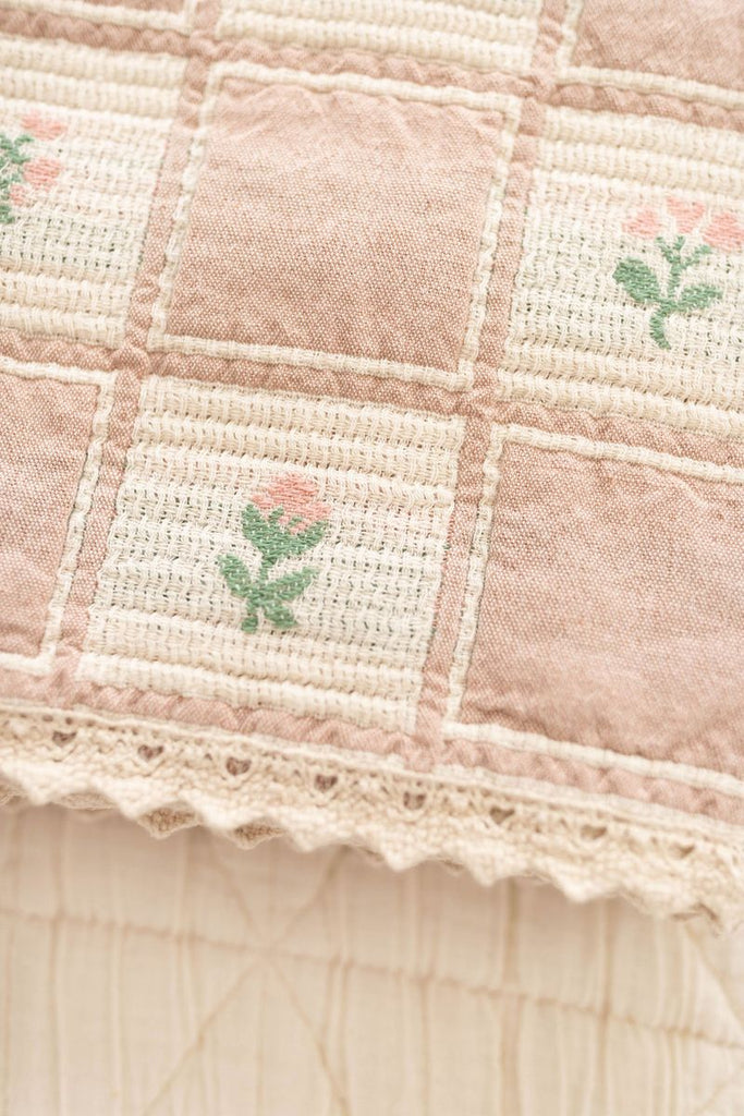 A close-up of the Organic Cotton Baby Blanket - Fleur shows its checkered pattern, pink embroidered flowers, and lace-trimmed edge. Made in Portugal from soft, vintage-style fabric.