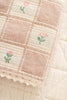 A close-up of the Organic Cotton Baby Blanket - Fleur, made in Portugal from soft organic cotton, shows its pink and cream checkered pattern, embroidered pink flowers, and lace-trimmed edge resting on textured light fabric.