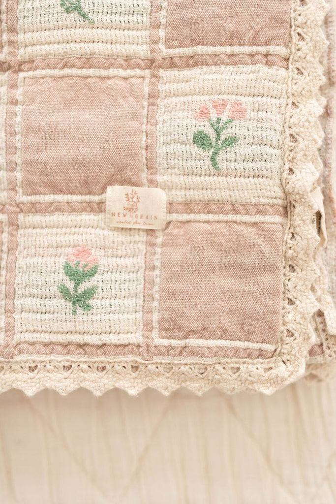 A close-up of the Organic Cotton Baby Blanket - Fleur shows its pink and cream checks, delicate embroidered flowers, lace trim, and a "NEVERFAIN" label. Made in Portugal from organic cotton.