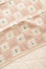 The Organic Cotton Baby Blanket - Fleur, made in Portugal, features a soft pink and cream patchwork design with small embroidered flowers and a delicate lace trim. It lies atop a quilted cream-colored surface.