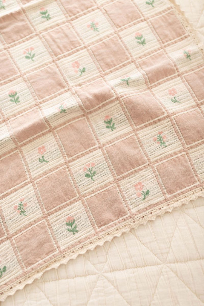 The Organic Cotton Baby Blanket - Fleur, made in Portugal, features a soft pink and cream patchwork design with small embroidered flowers and a delicate lace trim. It lies atop a quilted cream-colored surface.