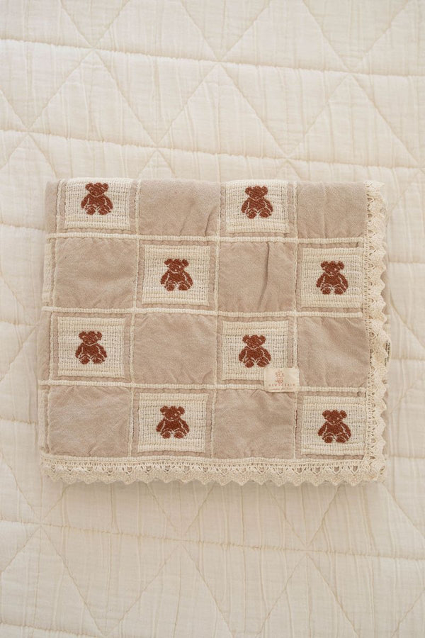 The Organic Cotton Baby Blanket - Teddy is a beige, quilted blanket with lace trim and a grid pattern featuring brown teddy bears, neatly folded on a cream quilt. Made in Portugal from soft, high-quality organic cotton.