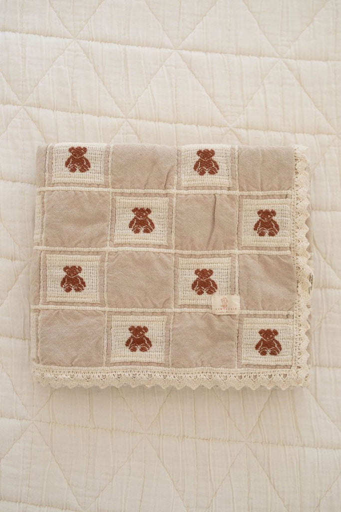 The Organic Cotton Baby Blanket - Teddy is a beige, quilted blanket with lace trim and a grid pattern featuring brown teddy bears, neatly folded on a cream quilt. Made in Portugal from soft, high-quality organic cotton.