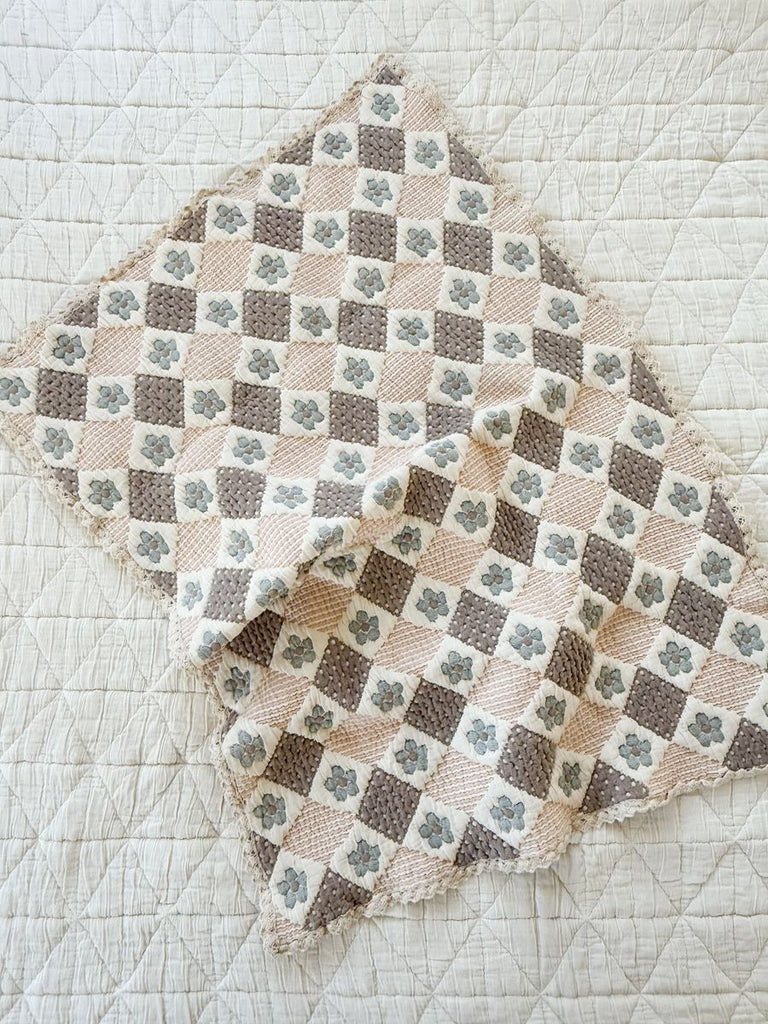 The Organic Cotton Baby Blanket - Prairie features a handmade checkered pattern in cream, beige, brown, and blue-gray with some blue-gray floral designs—ideal as a cozy baby blanket on any bedspread.