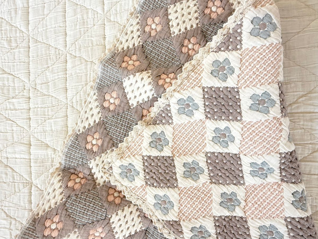 A close-up of the Organic Cotton Baby Blanket - Prairie, showcasing its pastel patchwork pattern with blue and pink flowers, textured stitching, and layered over a simple cream quilted background.