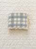 The Organic Cotton Baby Blanket - French Plaid, Blue features a blue and white plaid pattern with lace trim. Made from soft organic cotton in Portugal, it rests on an off-white quilted surface with a diamond design.