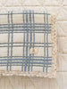 A folded Organic Cotton Baby Blanket - French Plaid, Blue with lace trim rests on a quilted cream bedspread. A sewn “EVERMAIN” label featuring a tree logo highlights its quality and made in Portugal craftsmanship.
