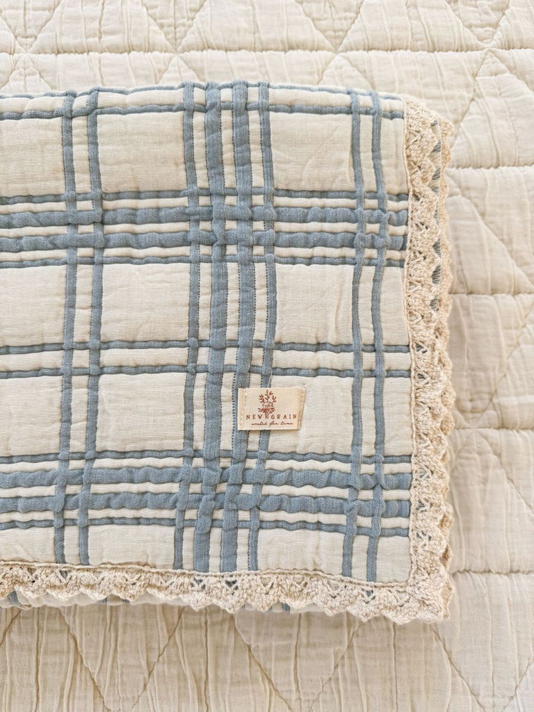 A folded Organic Cotton Baby Blanket - French Plaid, Blue with lace trim rests on a quilted cream bedspread. A sewn “EVERMAIN” label featuring a tree logo highlights its quality and made in Portugal craftsmanship.