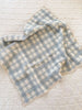 The Organic Cotton Baby Blanket - French Plaid, Blue features a soft, crumpled texture and blue and white plaid design, resting atop a cream-colored quilted surface.