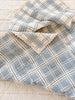 The Organic Cotton Baby Blanket - French Plaid, Blue features a soft blue and white plaid design with lace trim. Made in Portugal from organic cotton, this cozy blanket has a simple, handmade look and is shown partially folded on a light surface.