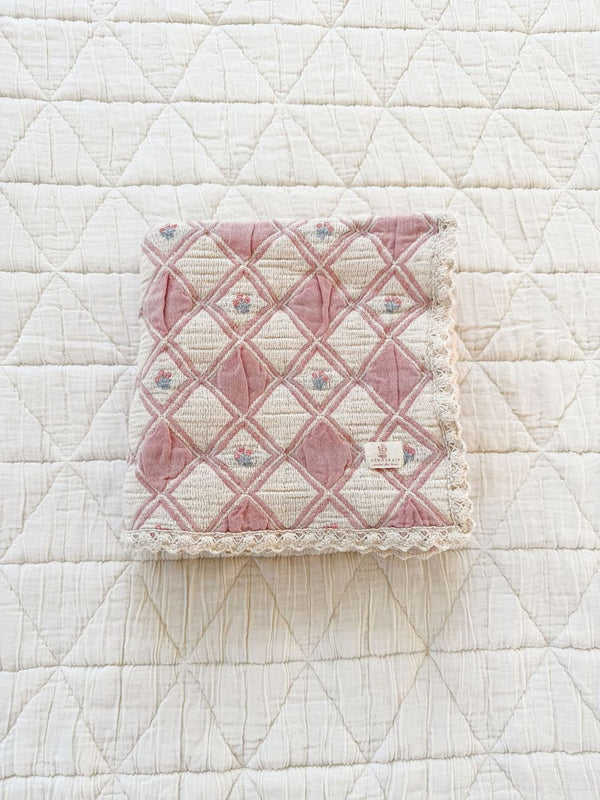 The Organic Cotton Baby Blanket - Bouquet features a folded pink and cream quilted design with a diamond pattern and lace edging, resting on a larger white quilted surface.
